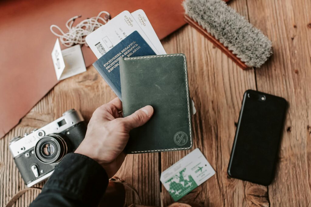 Close-up of a hand holding a passport with travel essentials including a camera, phone, and tickets on a wooden table.